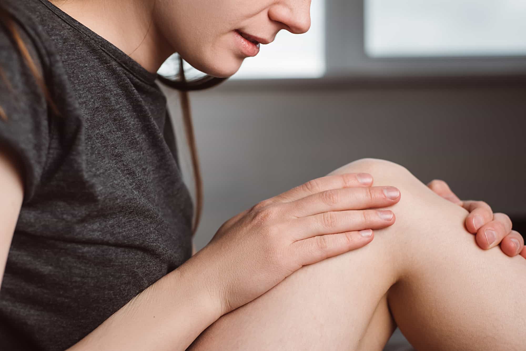 A young woman sitting on a bed at home is massaging and touching her knee with her hands due to joint pain