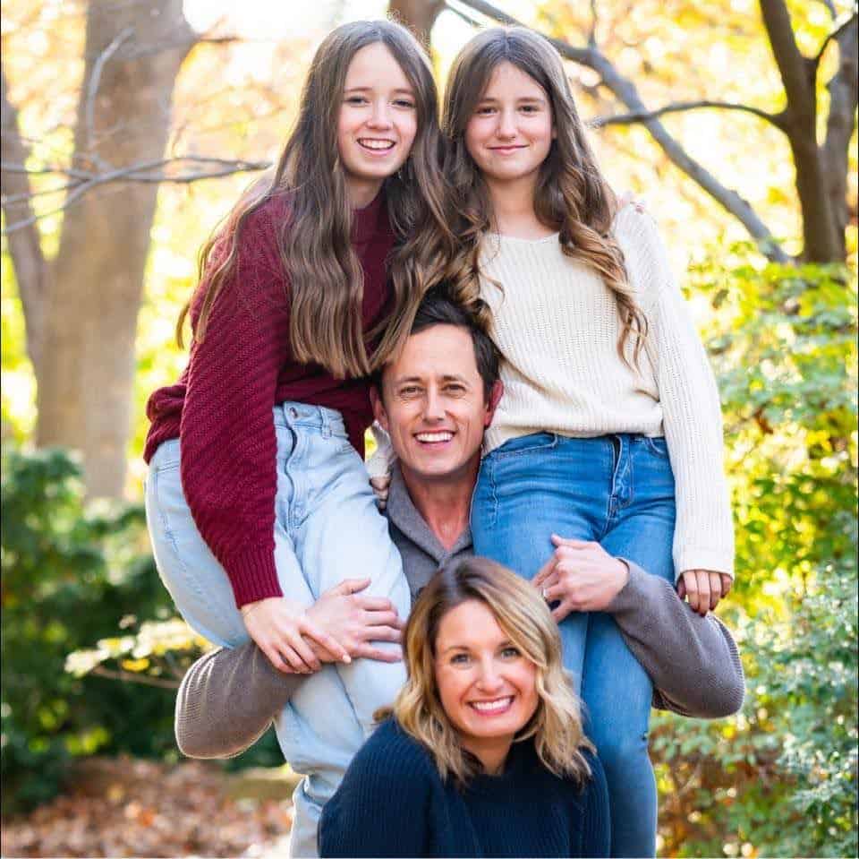 Family of four posing outdoors in autumn, with two young girls riding on parents' shoulders and everyone smiling for a portrait.